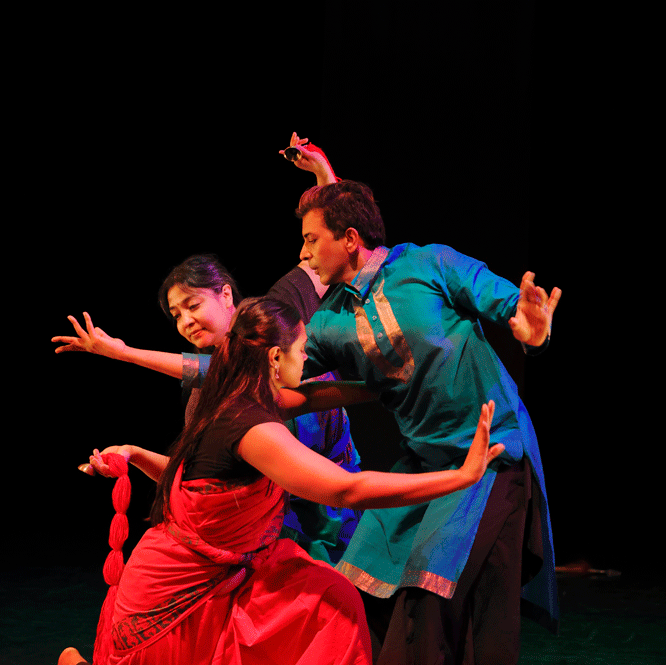 Three dance artists pose together on a dark stage. They are leaning over, into one another with their arms outstretched. They are wearing traditional Bharatanatyam clothing.