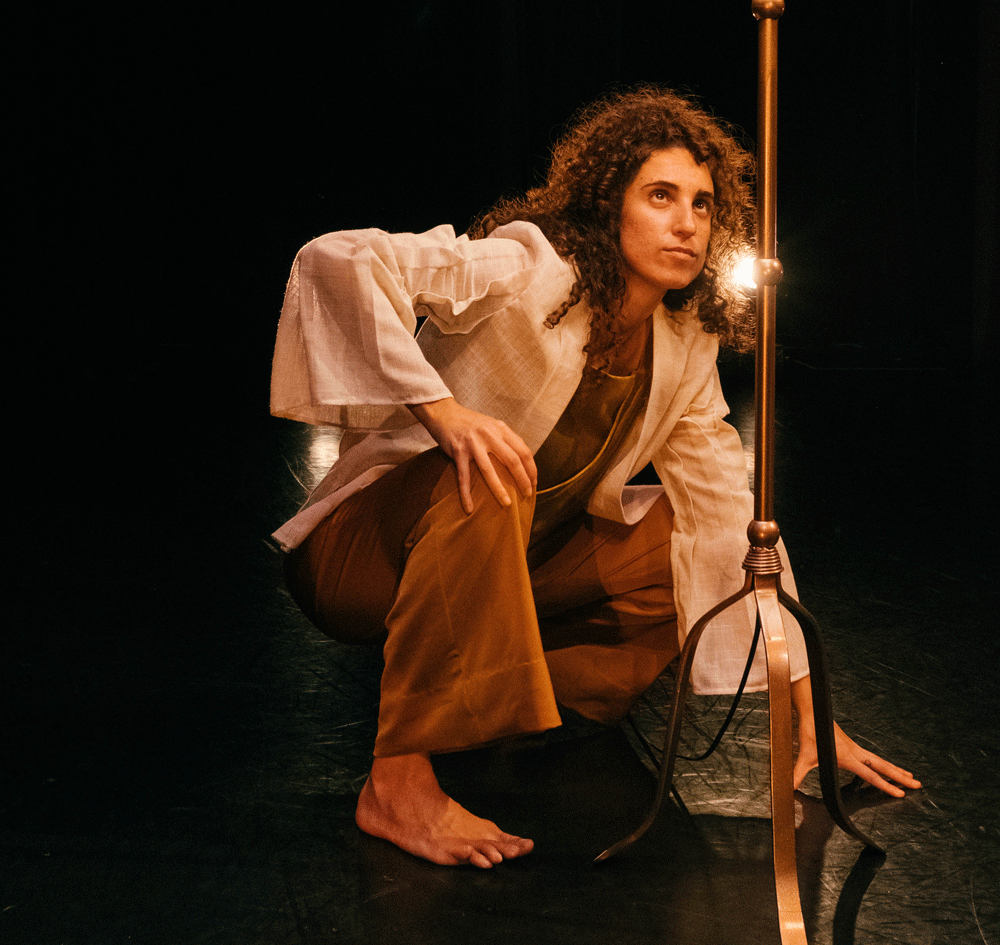 A dance artist crouches on the floor of a stage looking up at a floor lamp. They have brown curly hair and are wearing a brown jumpsuit and a white linen shirt over top.