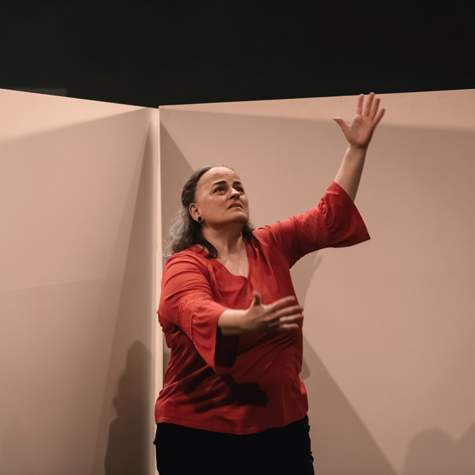 A dance artist stands in front of white backdrops. They are looking upwards and their arms are extended. They are wearing a red shirt.