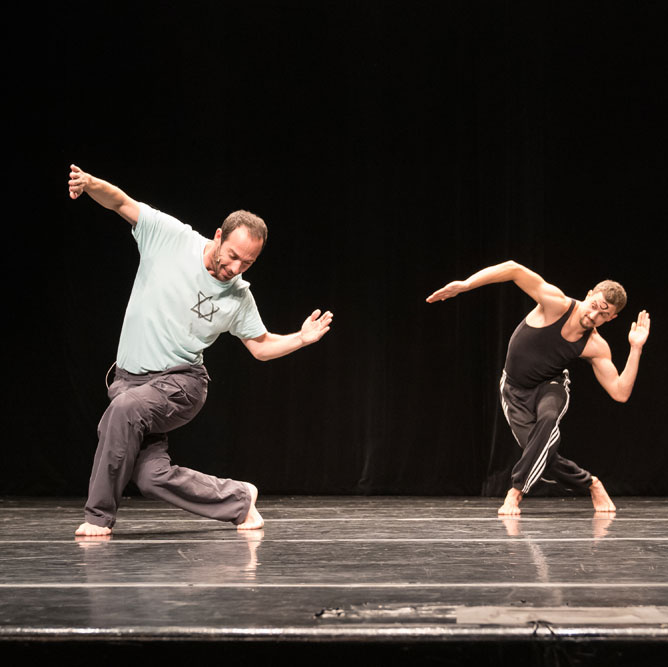 Two dance artists pose on a black shiny stage. They are wearing track pants, one has a black tank top on and the other has a blue tshirt with the Star of David in the left corner. They are both in mid curtsey with their arms extended. The one further back has a black almost circle painted on their forehead.