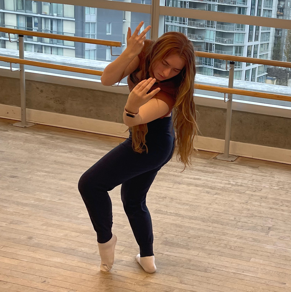 A dance artist with long red hair moves in a bright studio. They are bent to the left with their arms up and their eyes closed.