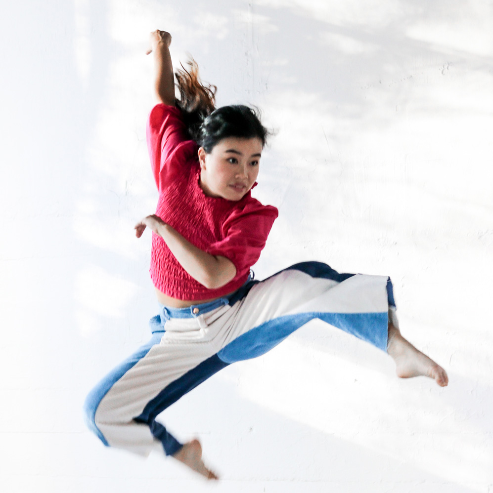 A dance artist jumps in the air. They are wearing a red sweater and blue and white striped pants. They have a dark ponytail and their right arm is extended behind them.