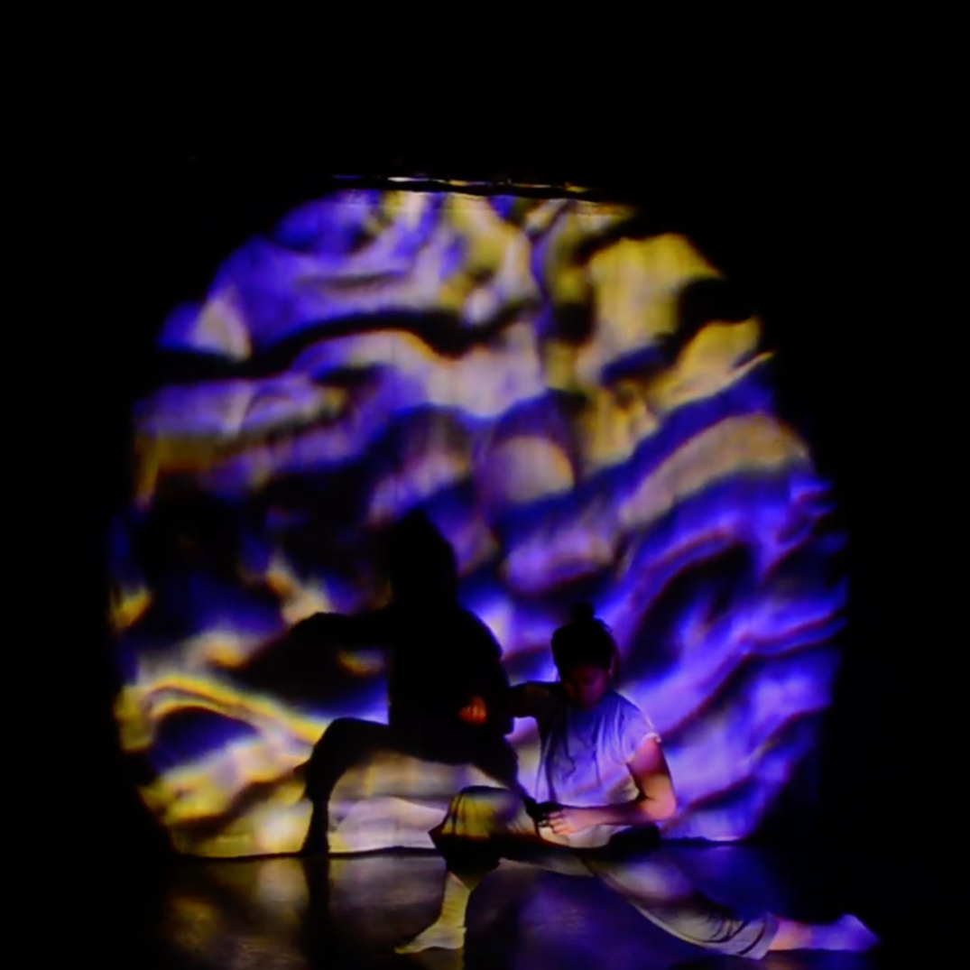 A dance artist poses in a low lunge on a dark stage. There is a textured purple and yellow spotlight over top